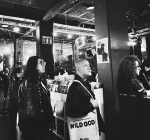 People browse vinyl records in a dimly lit record store; one person carries a tote bag labeled "WILD GOD.